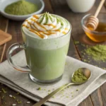 Glass mug of matcha latte topped with whipped cream, matcha drizzle, and honey on rustic wooden table