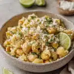 Elote pasta salad with ridged shells, cotija, cilantro, and lime in a speckled bowl.