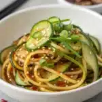 Spiralized cucumber salad with chili oil, green onions, and sesame seeds in a white bowl.