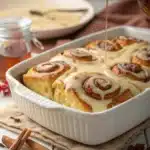 Maple syrup being drizzled over warm cinnamon roll casserole in a white baking dish.