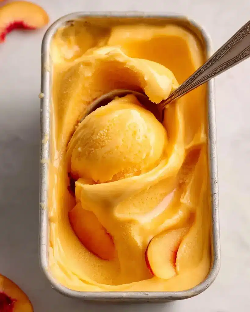 Creamy peach sorbet in a metal pan with a spoon and peach slices