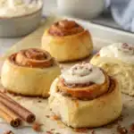 Freshly baked cinnamon rolls topped with creamy frosting, served warm with cinnamon sticks nearby.