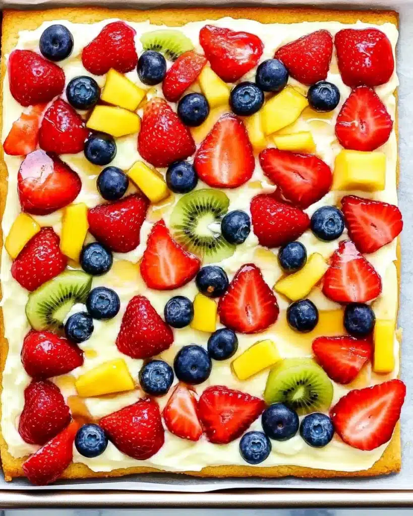 Uncut fruit pizza topped with strawberries, kiwi, mango, and blueberries on cream cheese frosting.