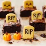 Halloween graveyard brownies with cookie tombstones and candy pumpkins on parchment paper.